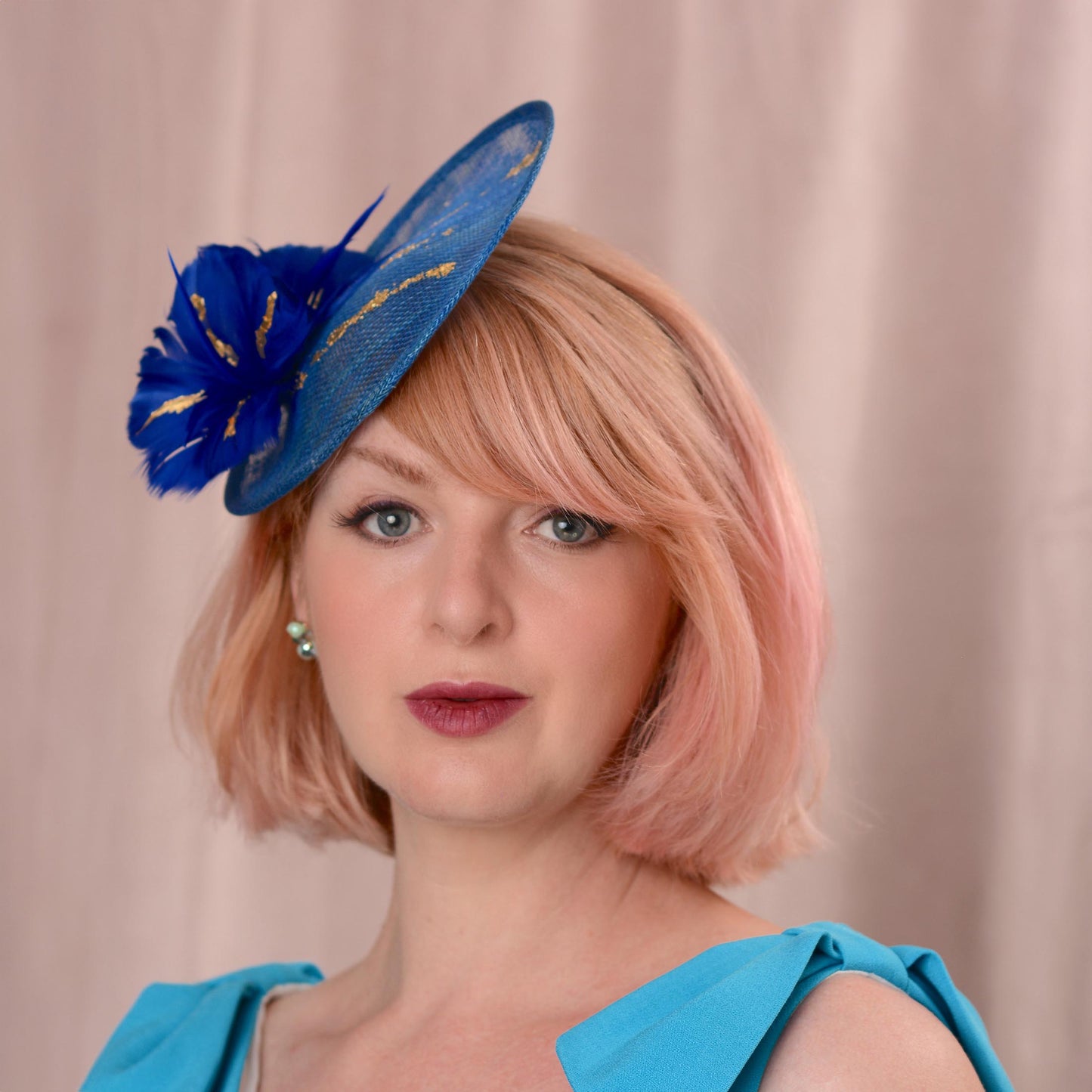 A woman with pink hair wearing a bright blue fascinator hat with gold accents and a matching blue top, photographed against a soft beige curtain background.