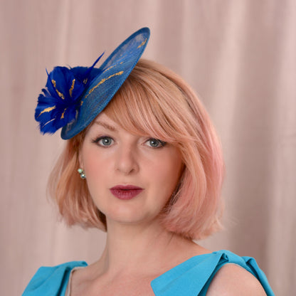 A woman with pink hair wearing a bright blue fascinator hat with gold accents and a matching blue top, photographed against a soft beige curtain background.