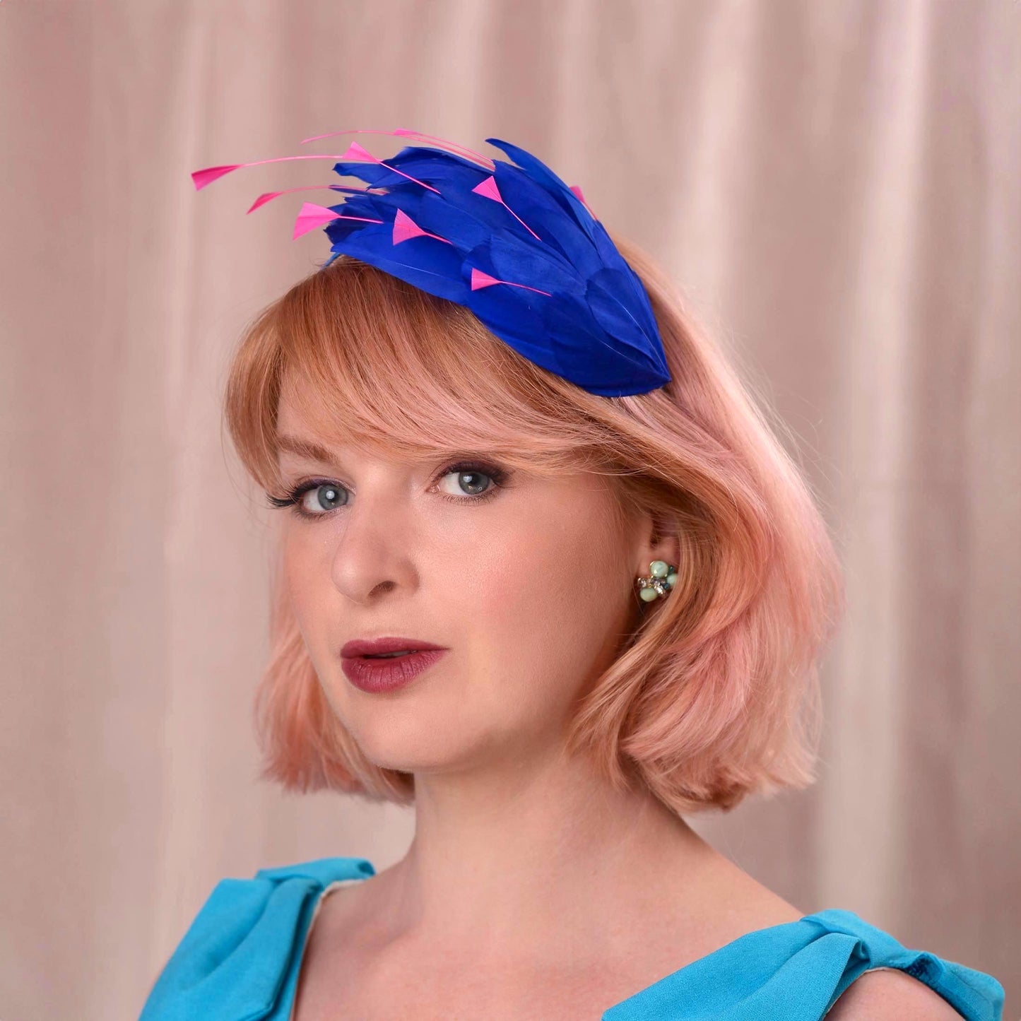 A woman with short pink hair wearing a bright royal blue feather fascinator hat with pink accents, paired with alight blu dress, set against a soft beige curtain background.