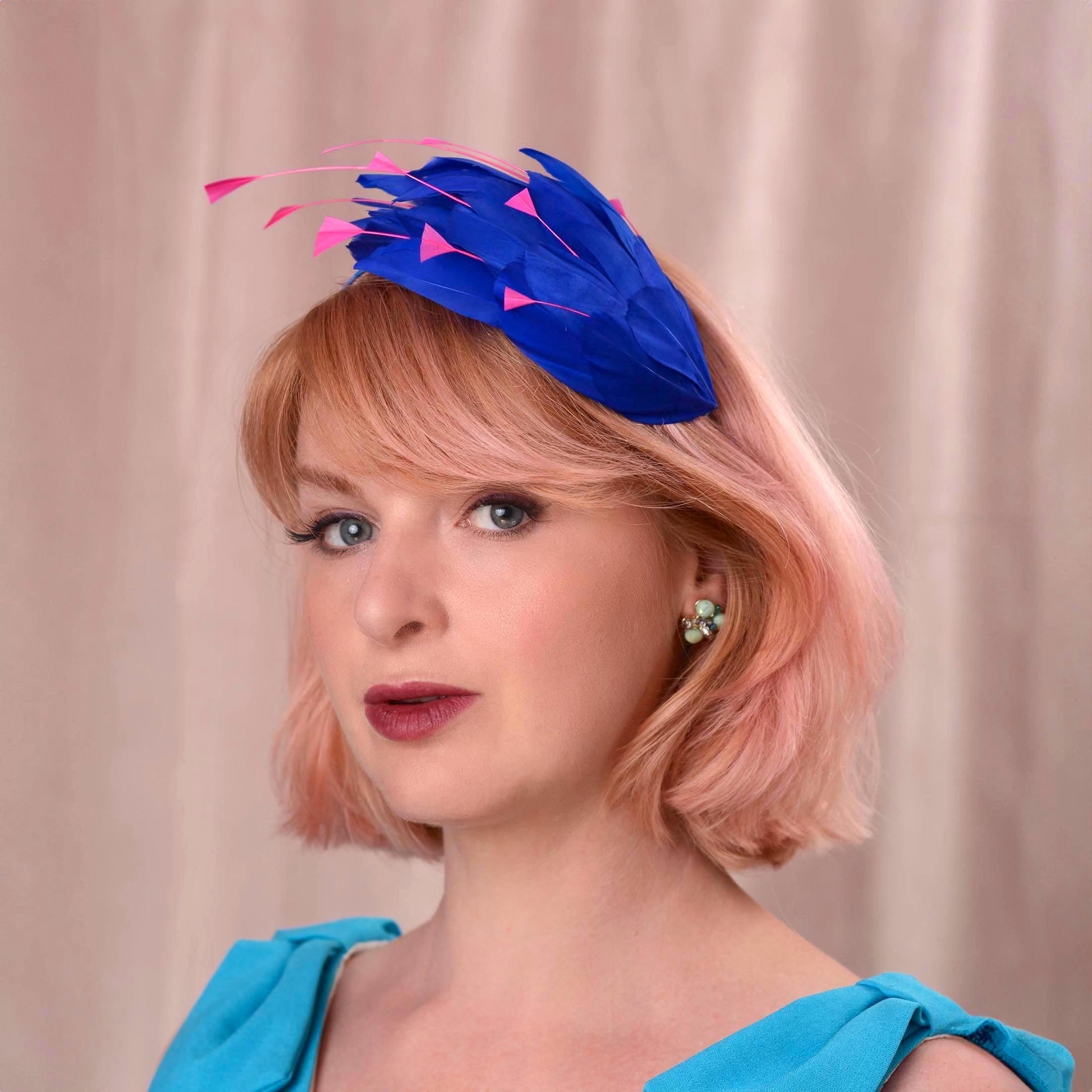 A woman with short pink hair wearing a bright royal blue feather fascinator hat with pink accents, paired with alight blu dress, set against a soft beige curtain background.
