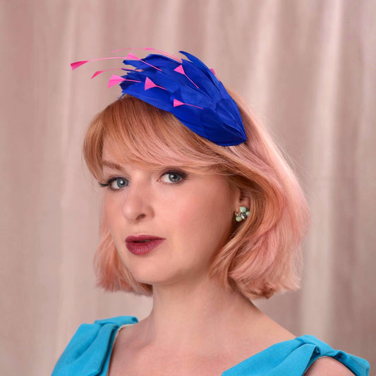 A woman with short pink hair wearing a bright royal blue feather fascinator hat with pink accents, paired with alight blu dress, set against a soft beige curtain background.