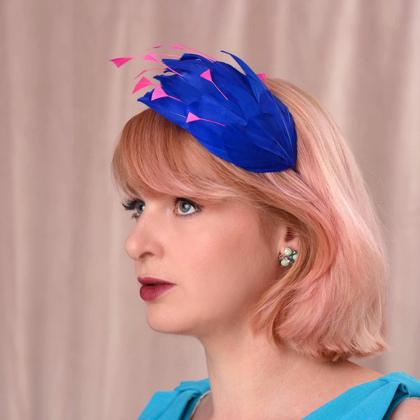 A woman with short pink hair wearing a bright royal blue fascinator headpiece with pink feather accents, shown in profile against a soft beige curtain background.