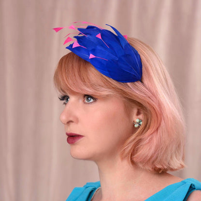 A woman with short pink hair wearing a bright royal blue fascinator headpiece with pink feather accents, shown in profile against a soft beige curtain background.