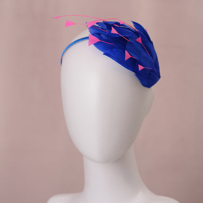 A royal blue fascinator headpiece with layered  shaped feathers and bright pink triangular  clipped feather accents, displayed on a white mannequin head against a soft dusty pink background.
