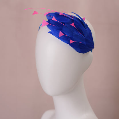 A royal blue feather fascinator headpiece with bright pink triangular feather accents, displayed on a white mannequin head against a soft pink background.