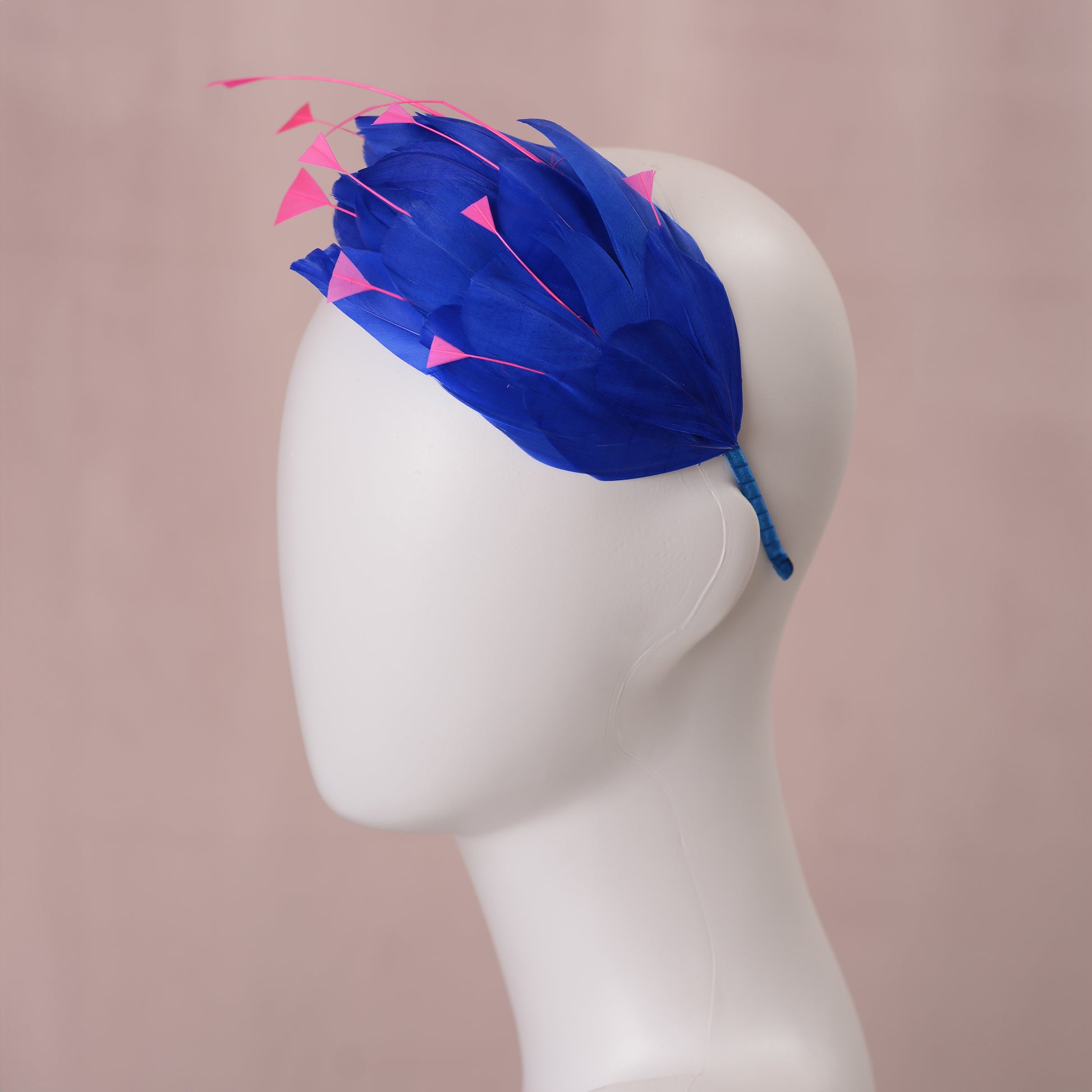 A royal blue feather fascinator headpiece with bright pink feather accents, displayed on a white mannequin head against a soft pink background.