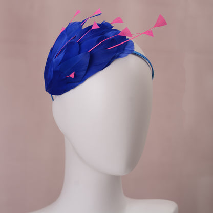 A royal blue feather fascinator headpiece with bright pink feather accents and a matching blue headband, displayed on a white mannequin head against a soft pink background.