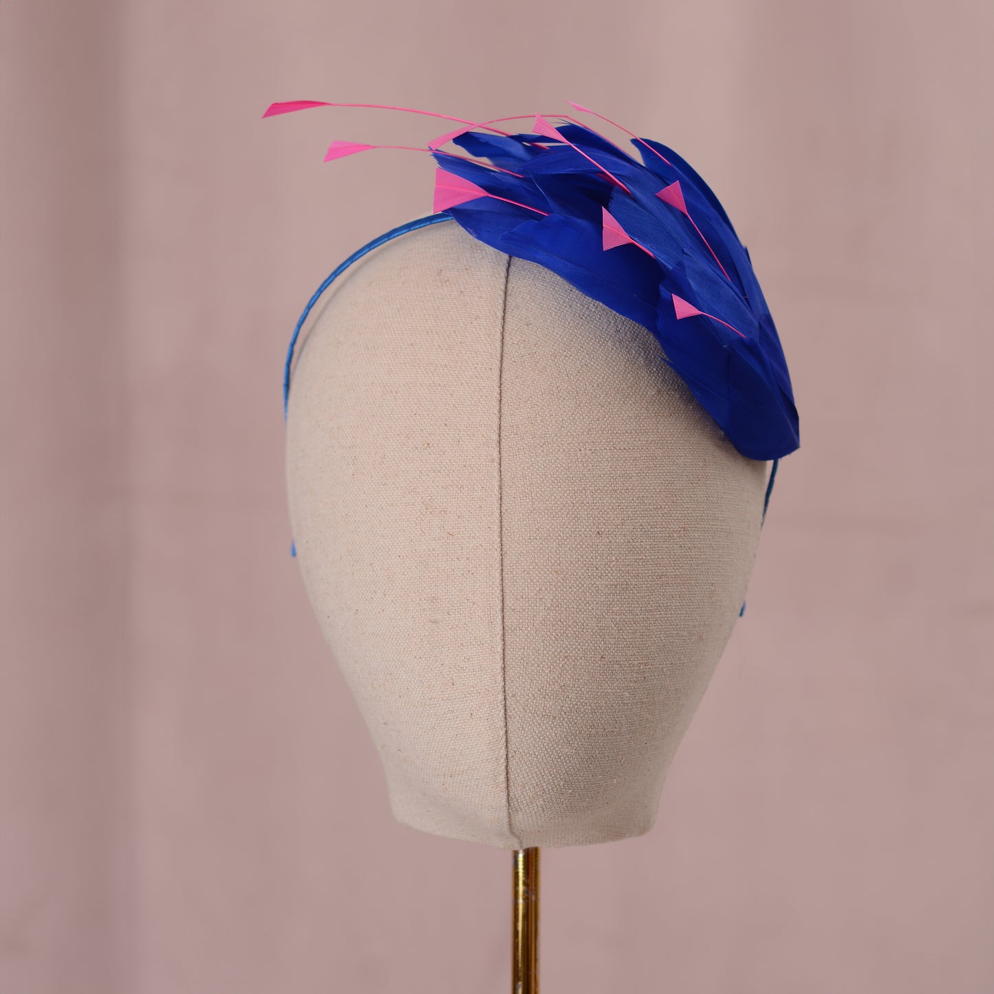 A blue and pink feather fascinator headpiece displayed on a beige fabric mannequin head against a soft pink background.