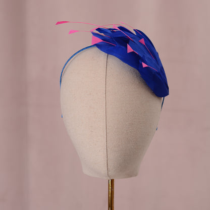 A blue and pink feather fascinator headpiece displayed on a beige fabric mannequin head against a soft pink background.