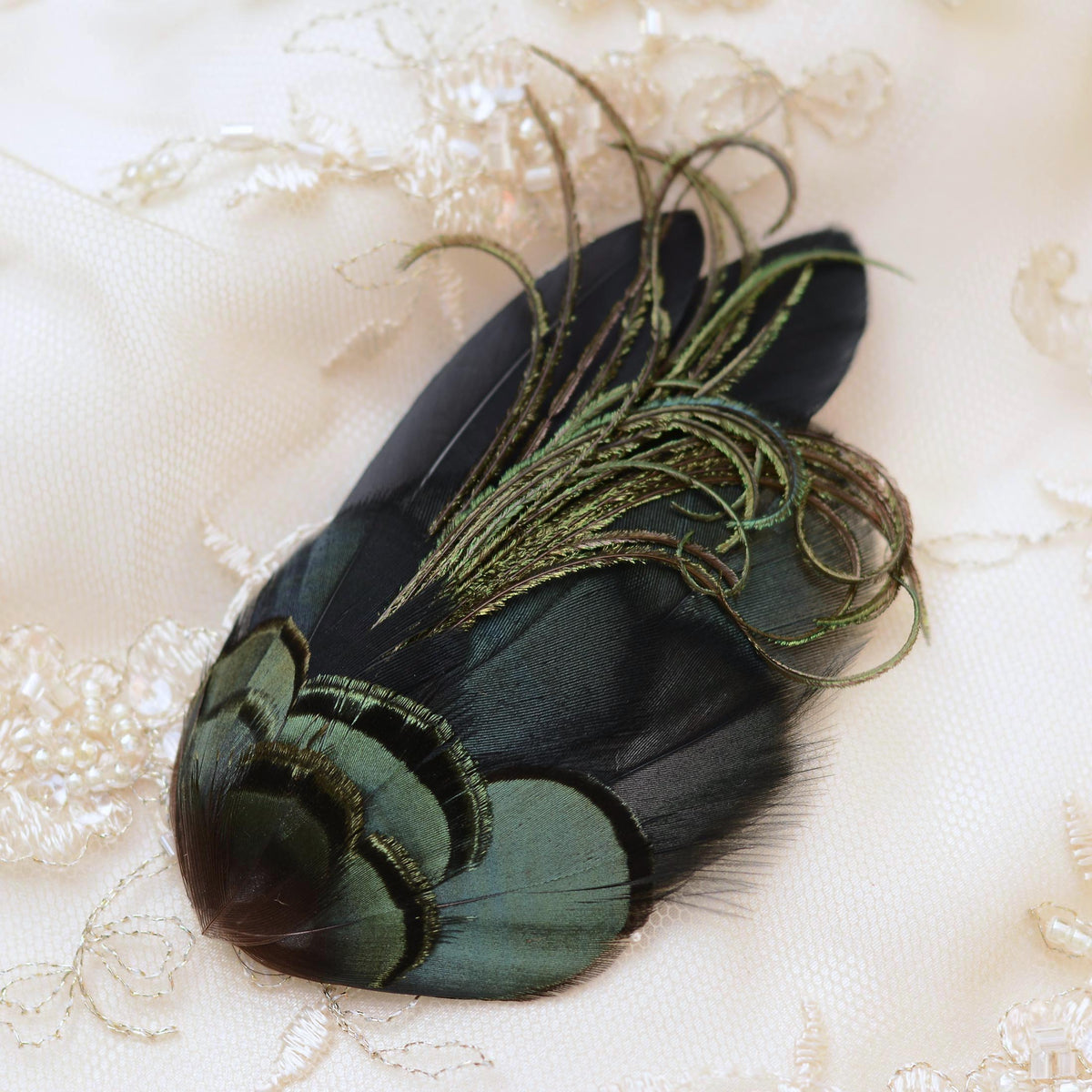 A decorative feather accessory with layered iridescent green and black feathers and long metallic olive-green plumes, placed on a cream lace fabric background.