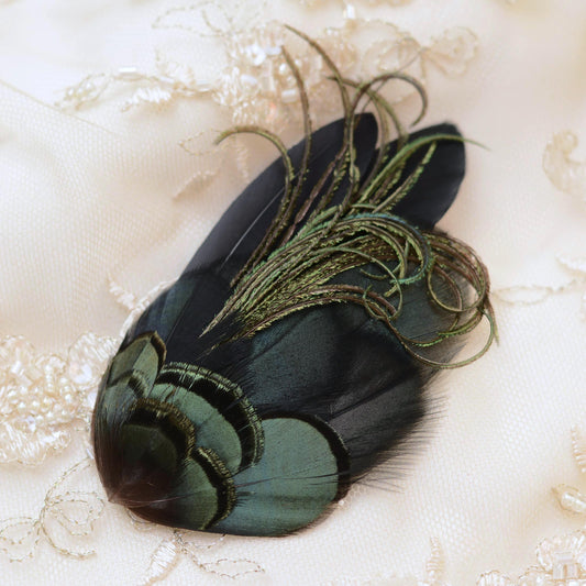 A decorative feather accessory with layered iridescent green and black feathers and long metallic olive-green plumes, placed on a cream lace fabric background.
