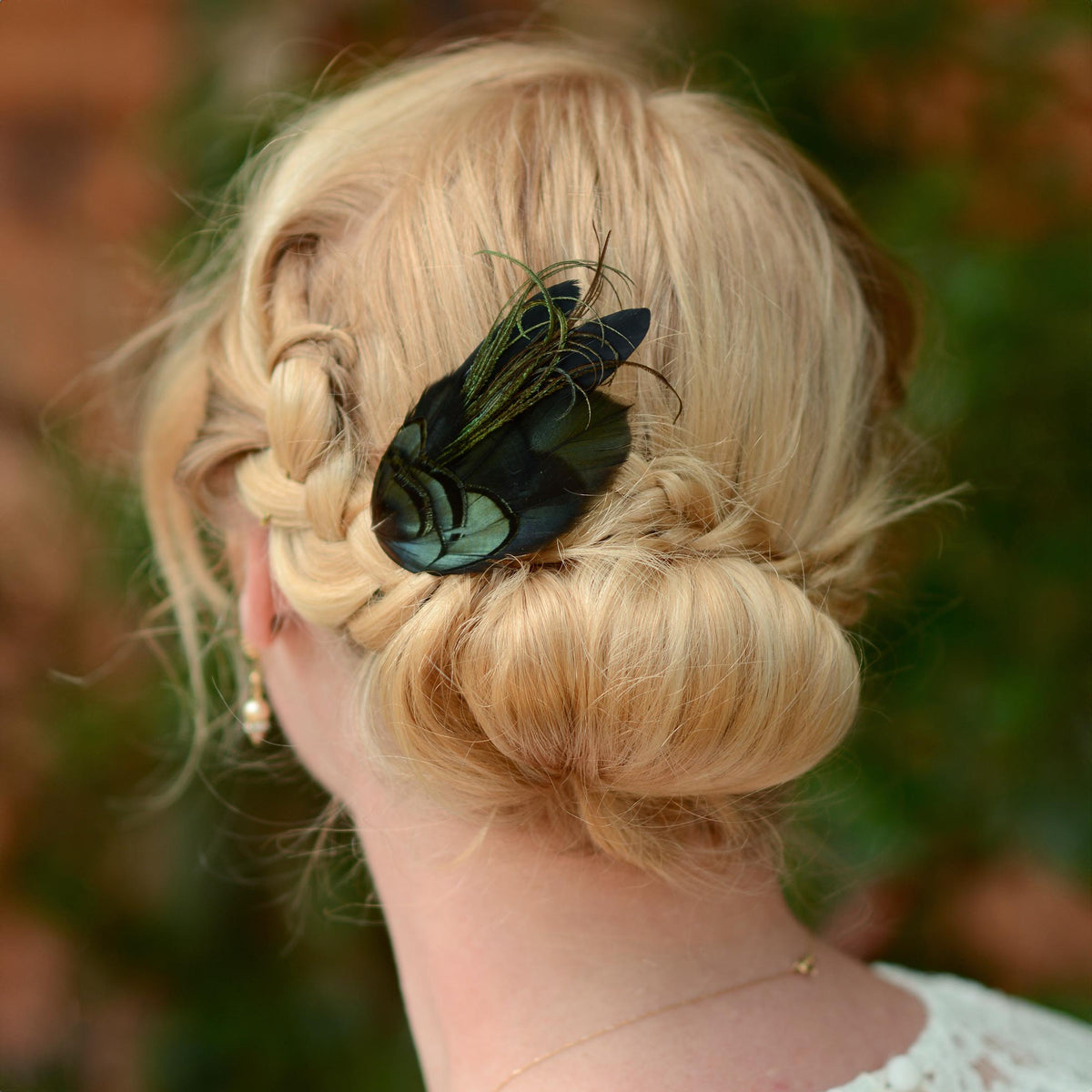 A blonde woman shown from the back wearing a decorative black and green feather hair accessory in her low bun, with a blurred outdoor background.