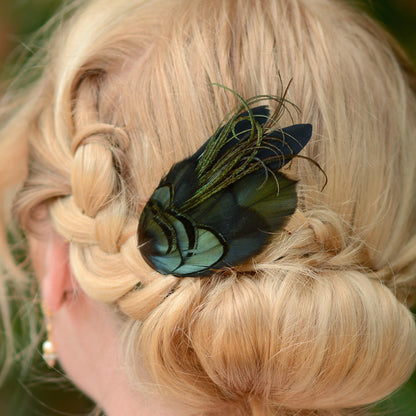 A close-up of a blonde woman's braided hair styled in a low bun, featuring a decorative hair accessory with iridescent blue-green feathers on black feathers with and fine curled accents.