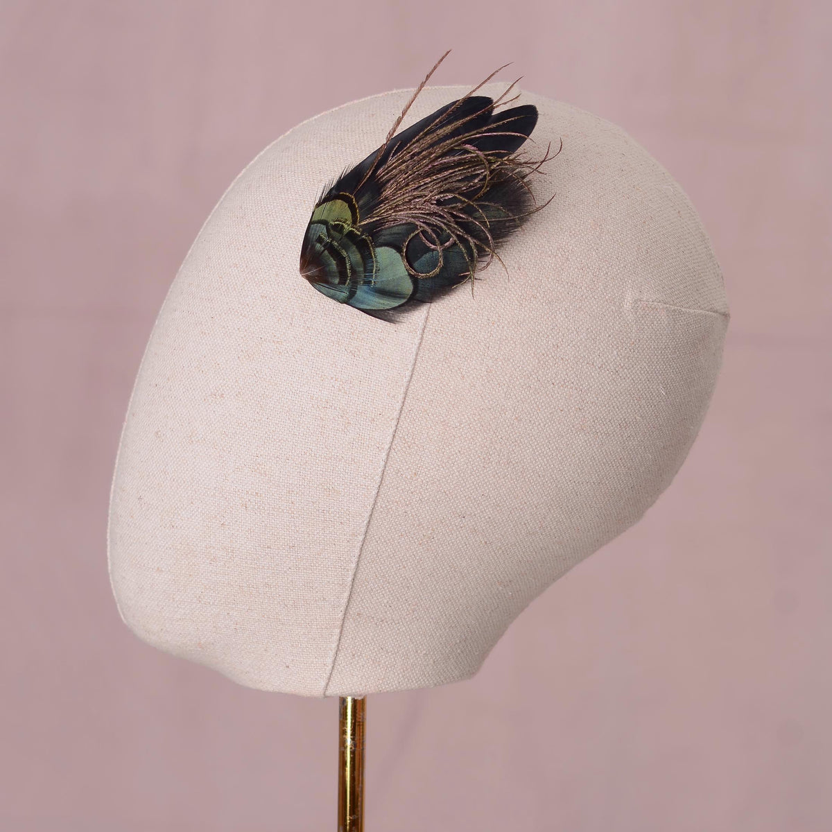 A decorative feather hair accessory with iridescent green and black feathers, displayed on a beige fabric mannequin head against a soft pink background.