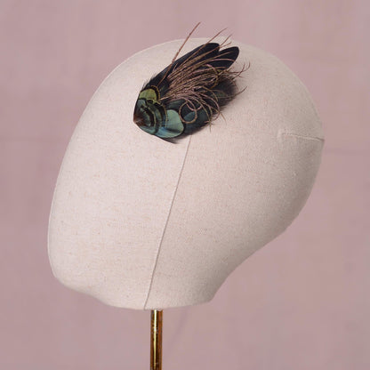 A decorative feather hair accessory with iridescent green and black feathers, displayed on a beige fabric mannequin head against a soft pink background.