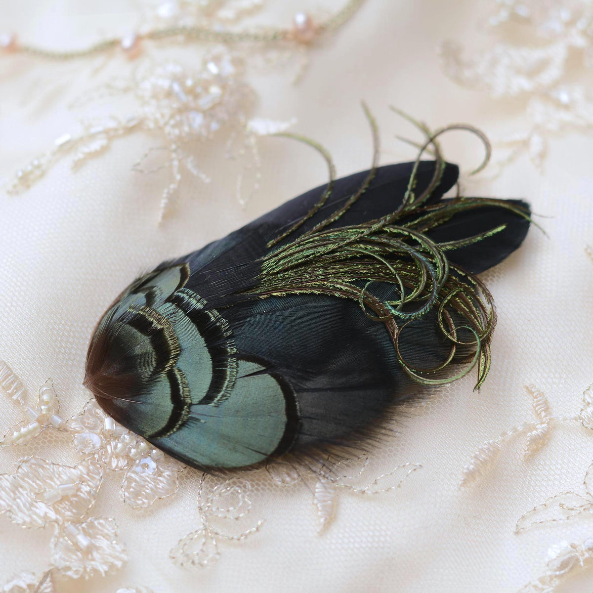 A decorative feather hair accessory with iridescent blue-green and black feathers, accented by shimmering metallic green curls, displayed on a cream embroidered fabric background.