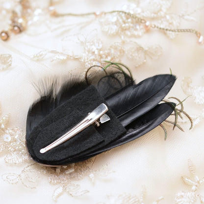 A black feather hair clip with a silver metal alligator-style clip attachment, displayed on a textured cream lace fabric background.