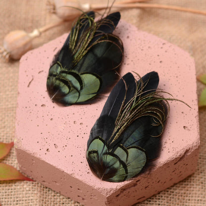 Two decorative feather clips or hair accessories with iridescent green and black feathers and purplish green delicate curled  plume strands, displayed on a pink textured stone block over a burlap surface.
