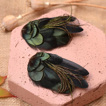 Two decorative feather clips or hair accessories with iridescent green and black feathers and purplish green delicate curled  plume strands, displayed on a pink textured stone block over a burlap surface.