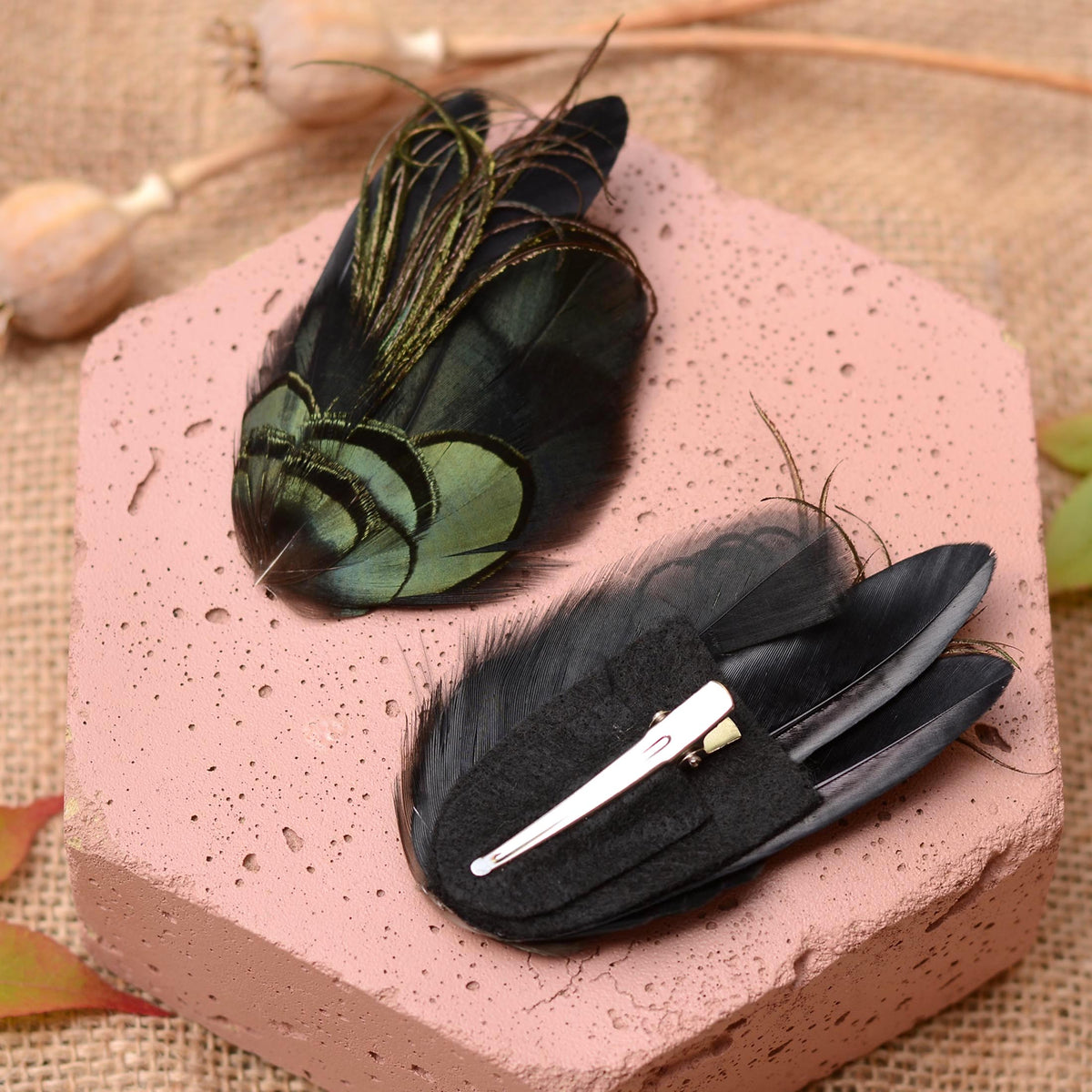 Two feather hair clips displayed on a pink hexagonal block. One clip features black feathers with iridescent green and gold tips, while the other is upside down to show the felt back and a silver metal alligator clip.