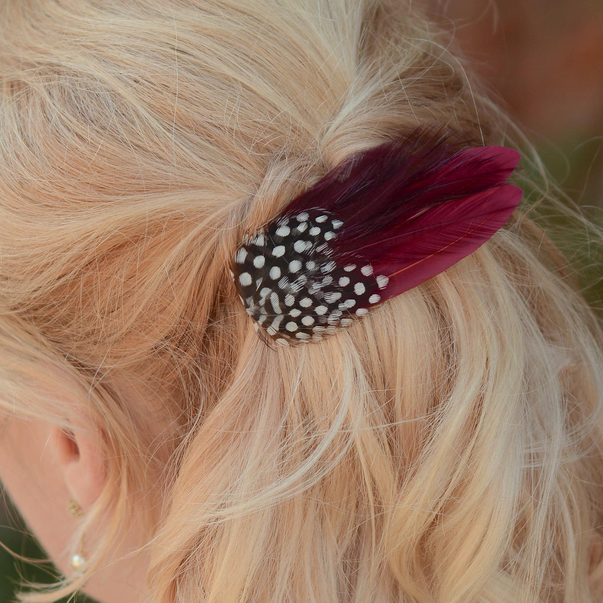 Close up of a woman with a blonde bob in a half up do adorned with a small feather hair accessory clip in burgundy and polka dot feathers. Perfect for accessorising a burgundy outfit or burgundy themed wedding.