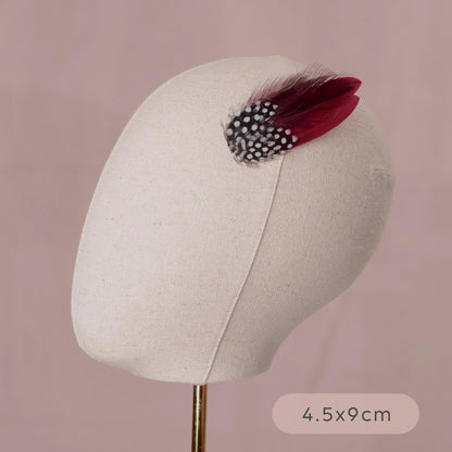 A cream-colored fabric mannequin head on a stand, decorated with a small feather accessory featuring a black and white polka-dot band and burgundy feathers.
