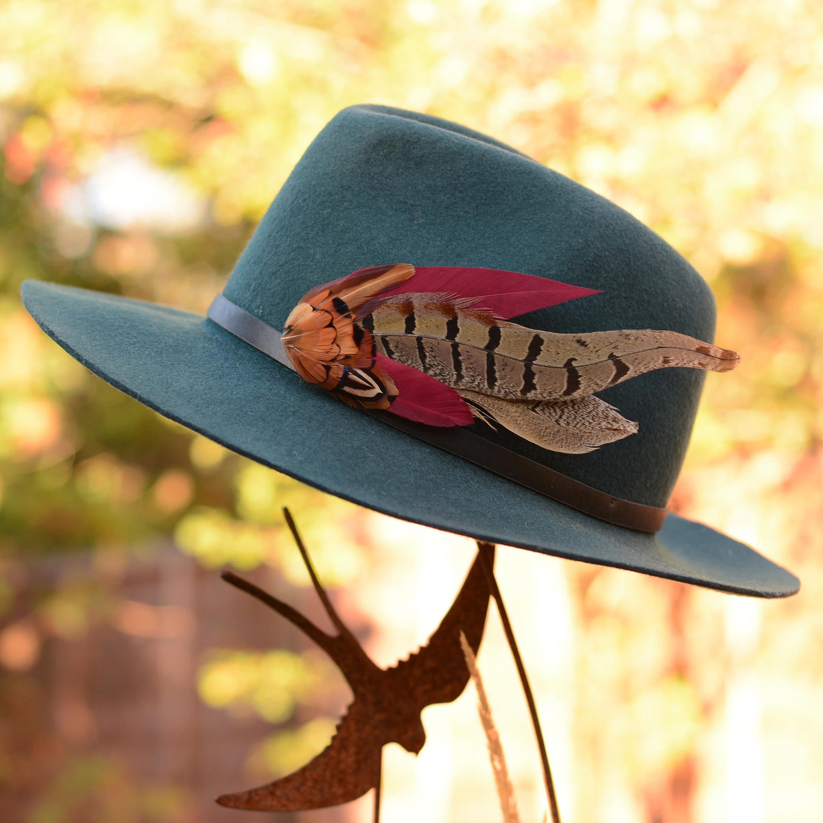 A statement burgundy and pheasant feather hat pin on a teal felt wide-brim fedora hat displayed on a metal stand outdoors.