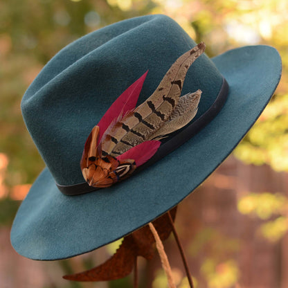 statement burgundy and pheasant feather hat pin perfect for cheltenham races and country events dispayed on a teal wool felt hat outside.