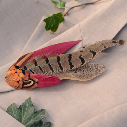 A decorative feather lapel pin with a large brown patterned pheasant tail feather,  burgundy feathers and copper-black feathers, styled on draped beige fabric with green leaves.