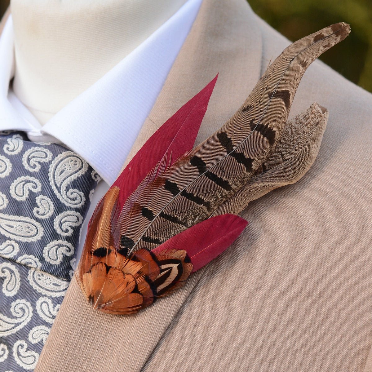 A decorative feather lapel pin with brown and copper patterned pheasant feathers and burgundy red accents, pinned to a beige suit jacket worn over a white shirt and a grey paisley tie.