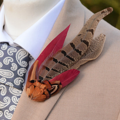 A decorative feather lapel pin with brown and copper patterned pheasant feathers and burgundy red accents, pinned to a beige suit jacket worn over a white shirt and a grey paisley tie.