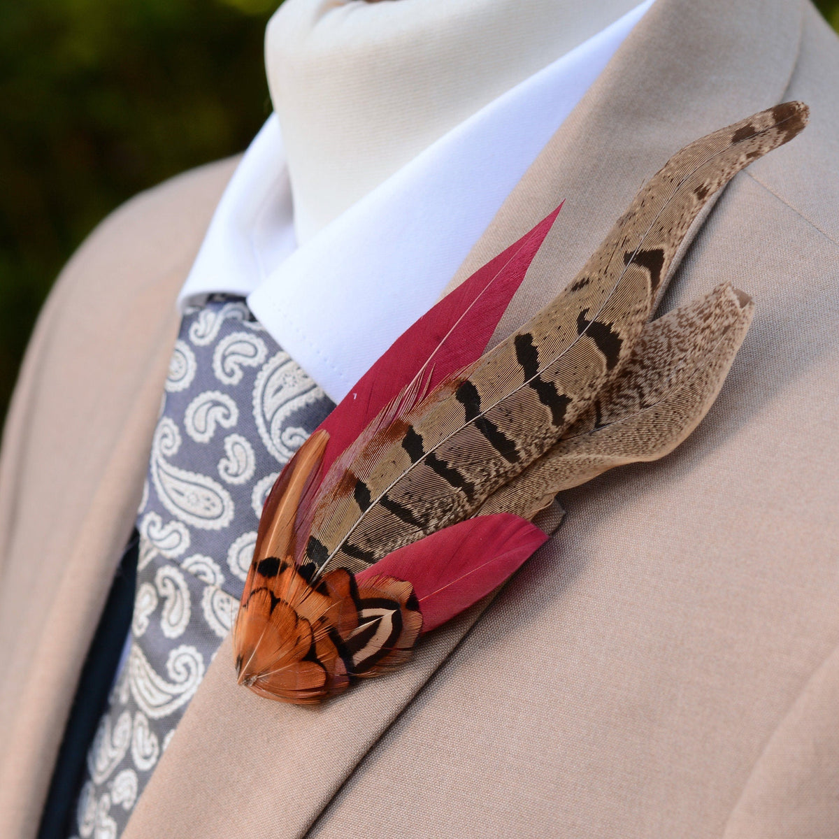 A close-up of a beige suit jacket on a mannequin or torso form, styled with a white dress shirt, grey paisley tie, and a large feathered lapel pin featuring brown patterned pheasant feathers and burgundy feathers.