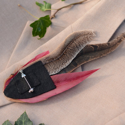 A feathered brooch or hat pin displayed on a beige fabric surface, featuring burgundy and brown feathers with a black felt base and a silver-toned pin attached.