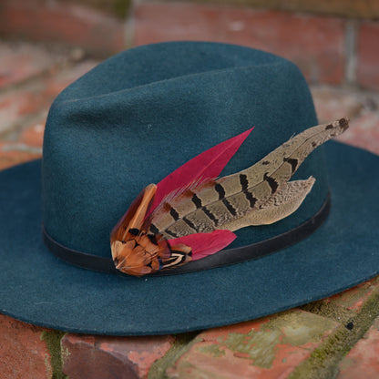 A dark teal felt hat with a black band and a decorative pheasant feather hat pin accent, resting on a brick surface.