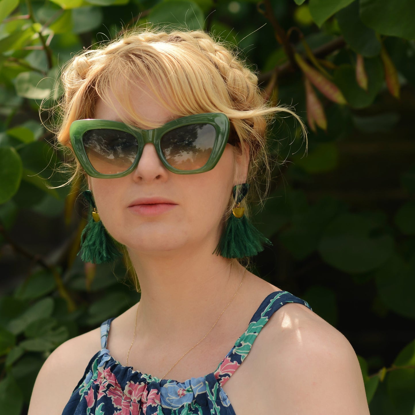 Woman wearing green sunglasses and green ostrich feather earrings with a floral dress against a leafy background
