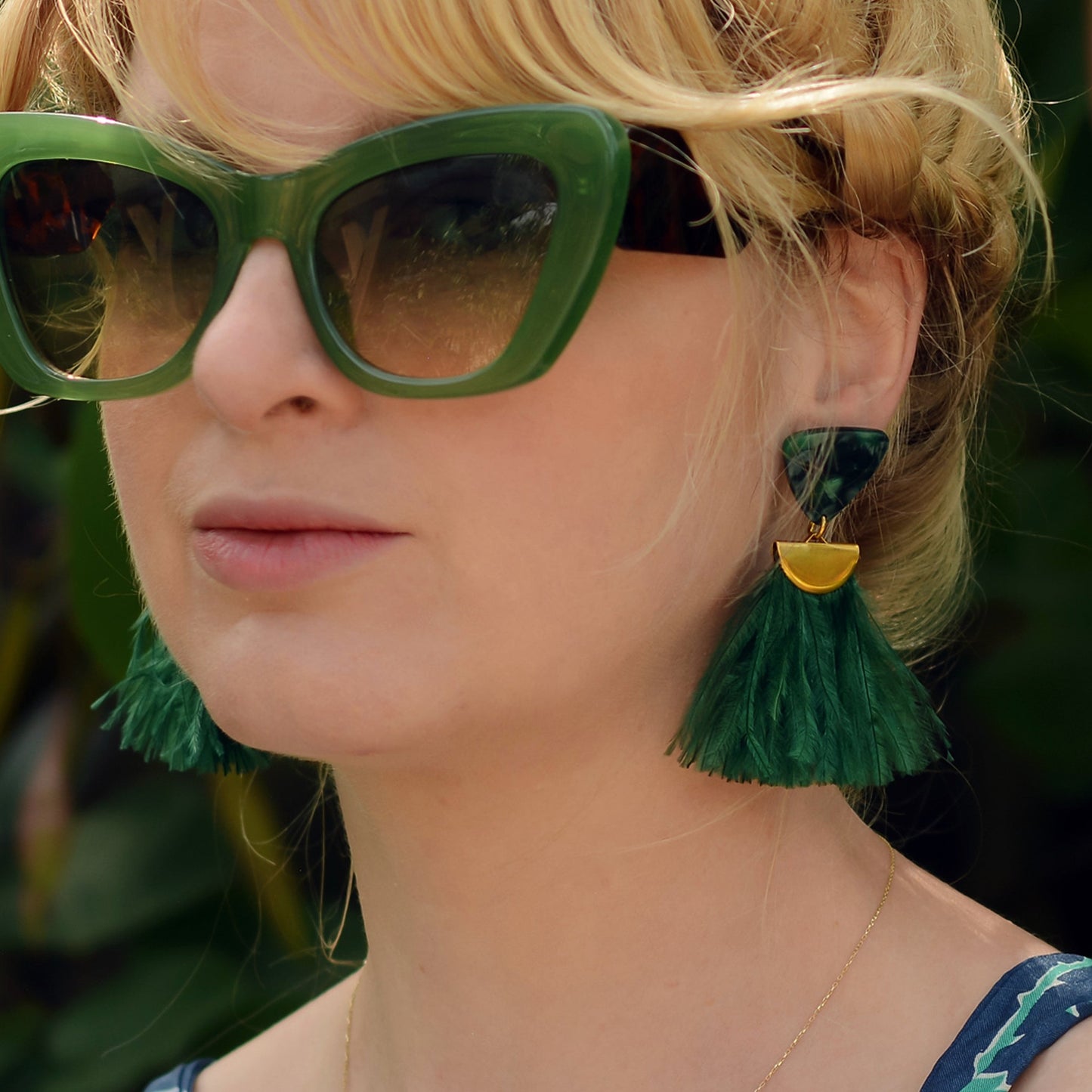 Close-up of a person wearing green sunglasses and green ostrich feather tassel earrings with a blurred background
