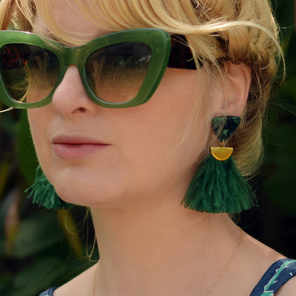 Close-up of a person wearing green sunglasses and green ostrich feather tassel earrings with a blurred background
