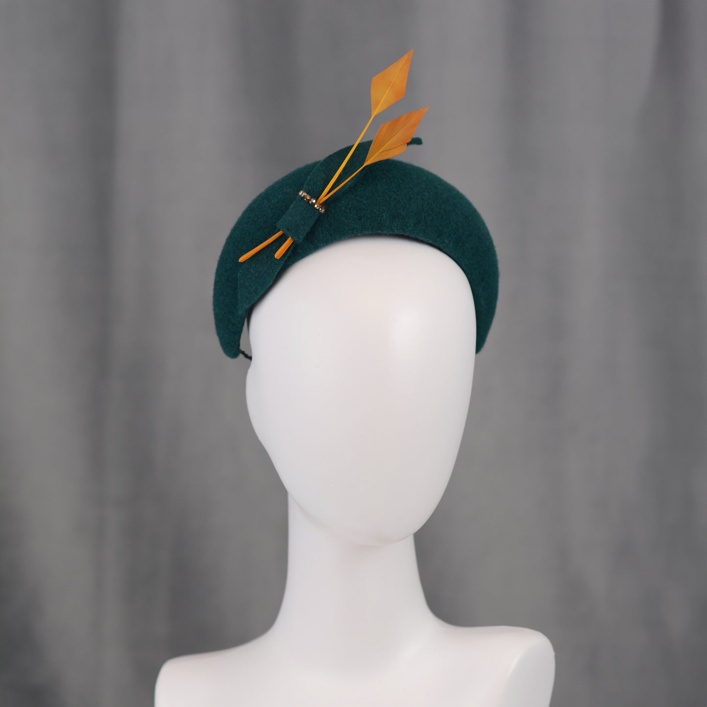 Teal green headband  fascinator with gold yellow feathers and bow on a mannequin with a grey background