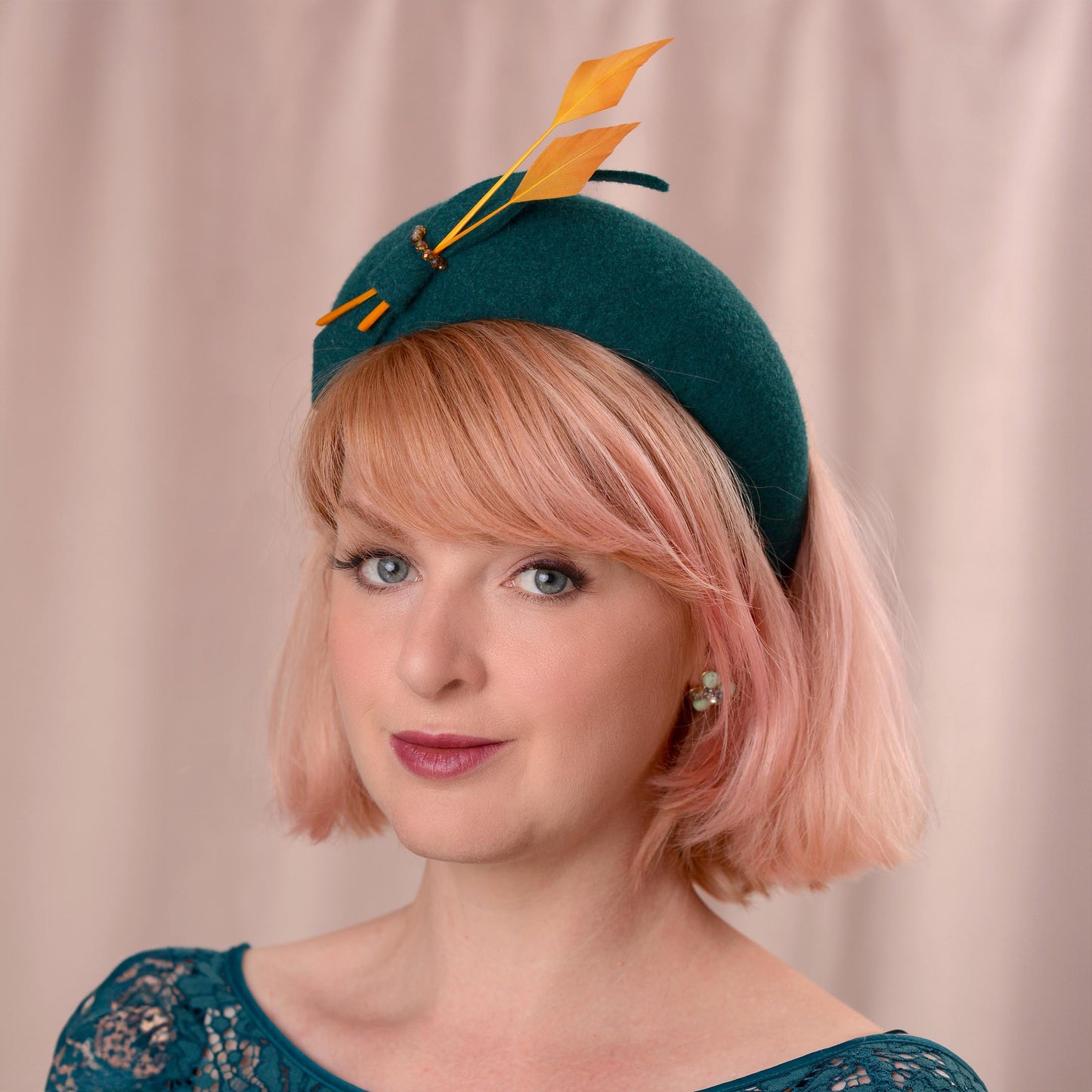 A woman with short pink hair models a dark teal felt wedding fascinator hat decorated with two gold yellow feathers and a small felt bow, set against a soft beige curtain background.