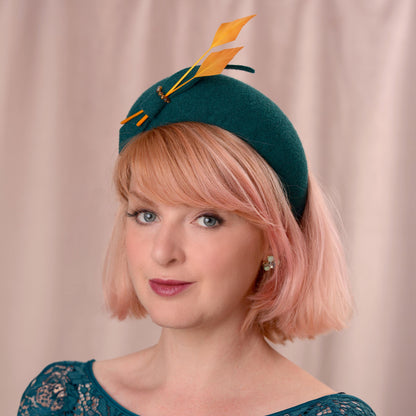 A woman with short pink hair models a dark teal felt wedding fascinator hat decorated with two gold yellow feathers and a small felt bow, set against a soft beige curtain background.