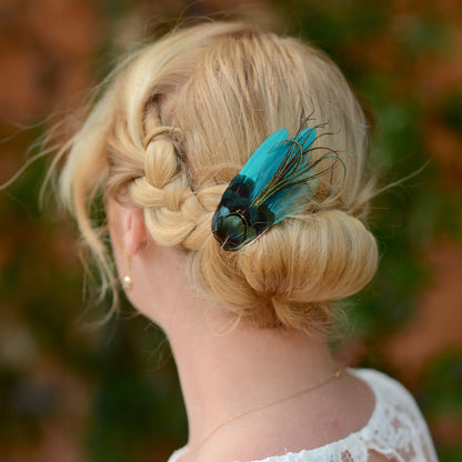 Blonde hair styled in an updo with a teal turquoise and green feather accessory against a blurred natural background