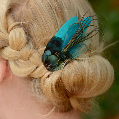 Blonde hair styled in an updo with a teal turquoise and green feather accessory against a blurred natural background