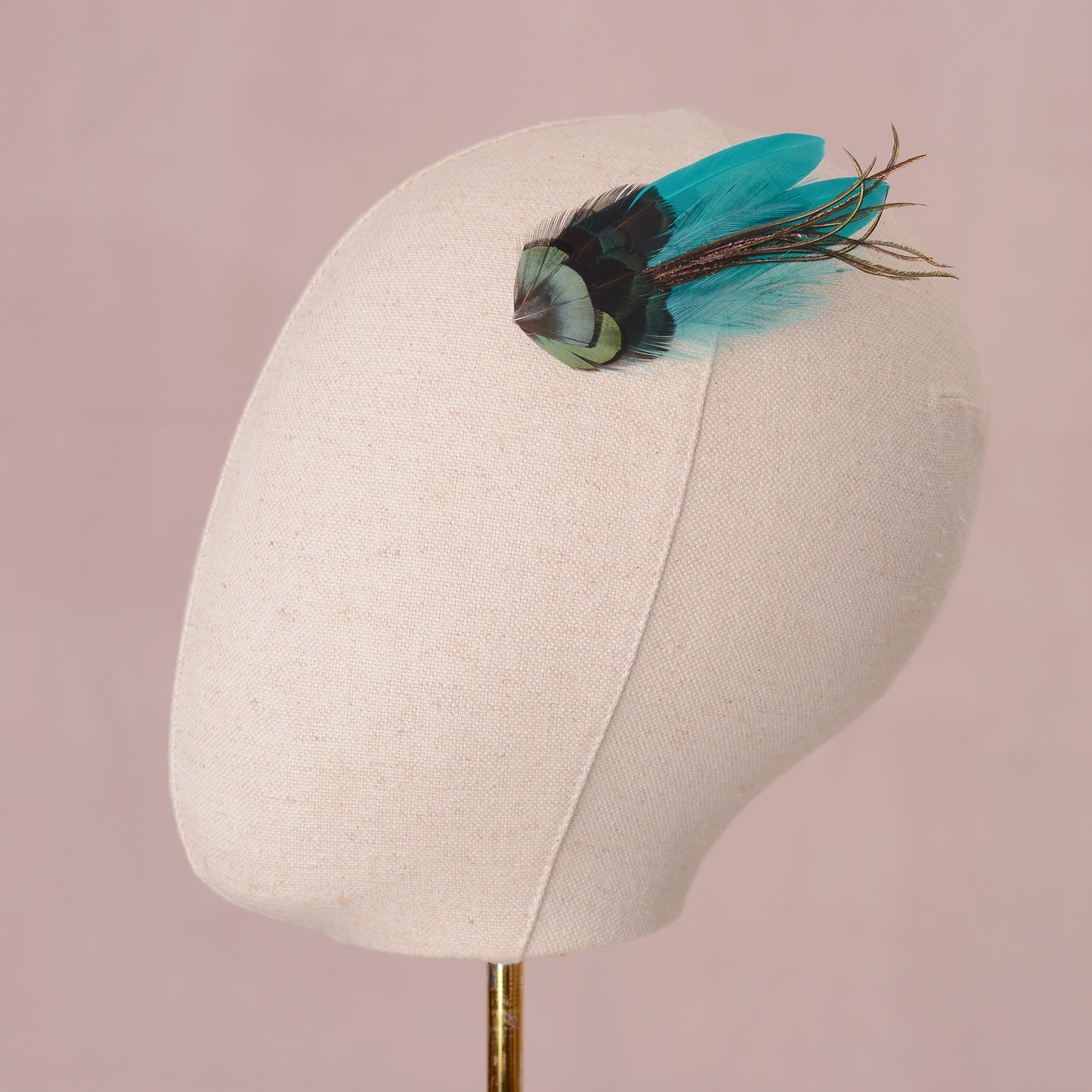 Mannequin head with a decorative teal, turquoise and green feather hairpiece on a pink background