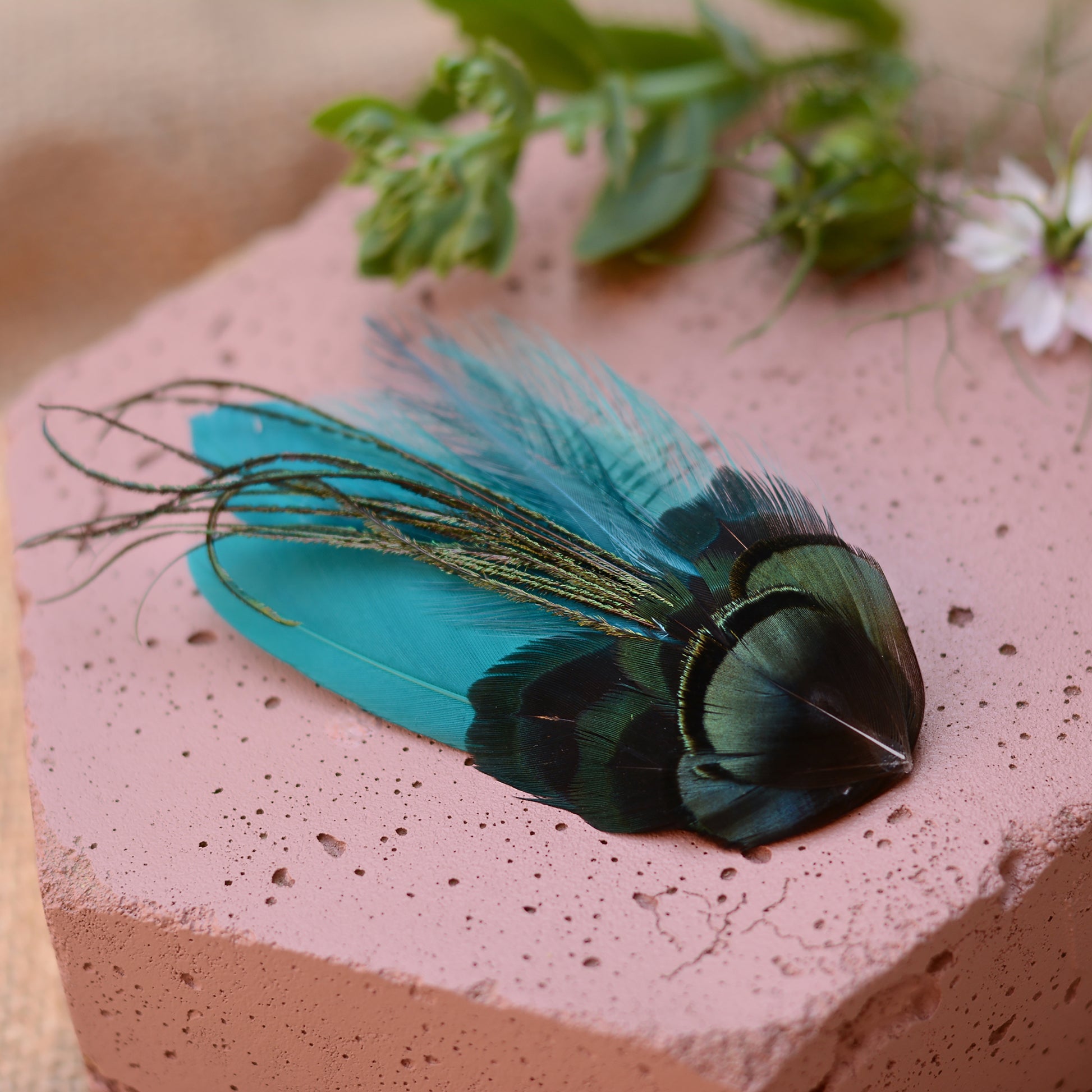 teal turquoise feather hair clip with iridescent green feathers on a pink stone background with flowers