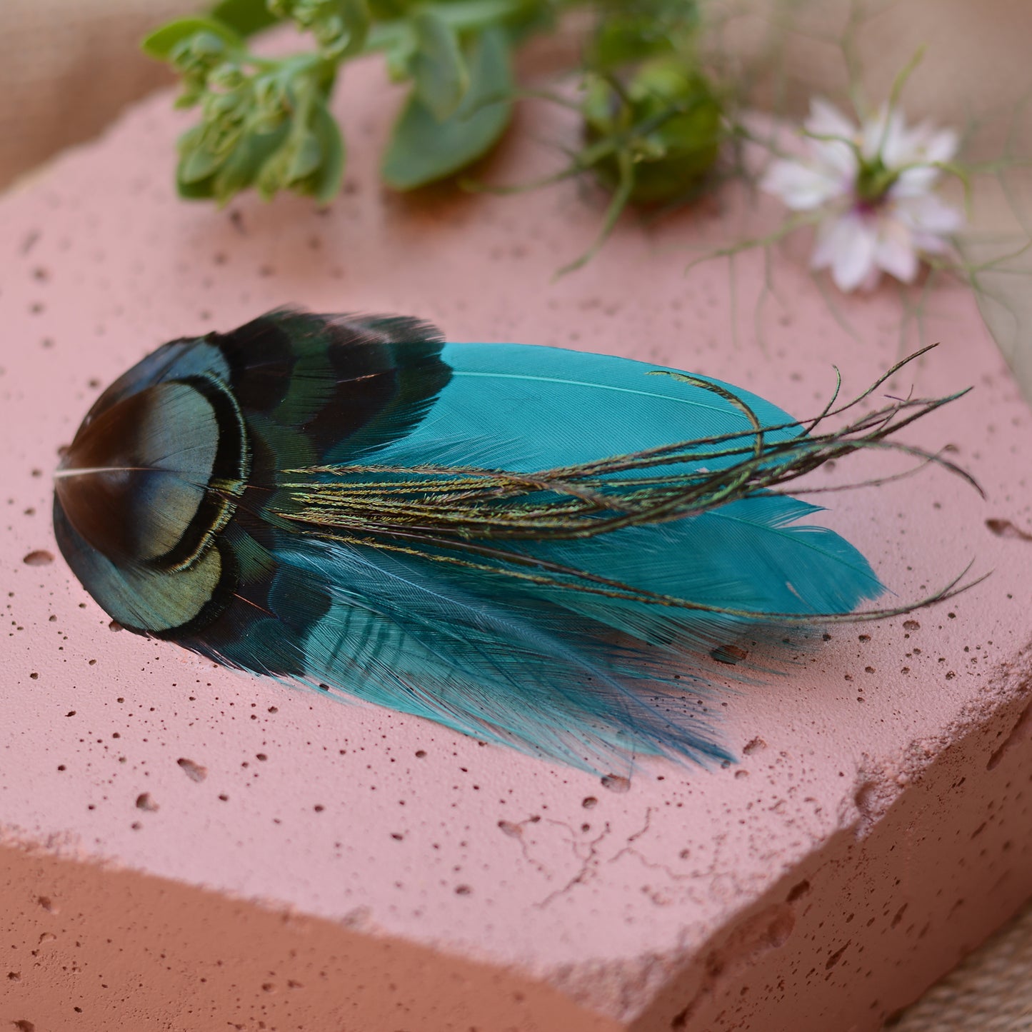 teal feather hair clip with iridescent green feathers on a pink stone background with flowers