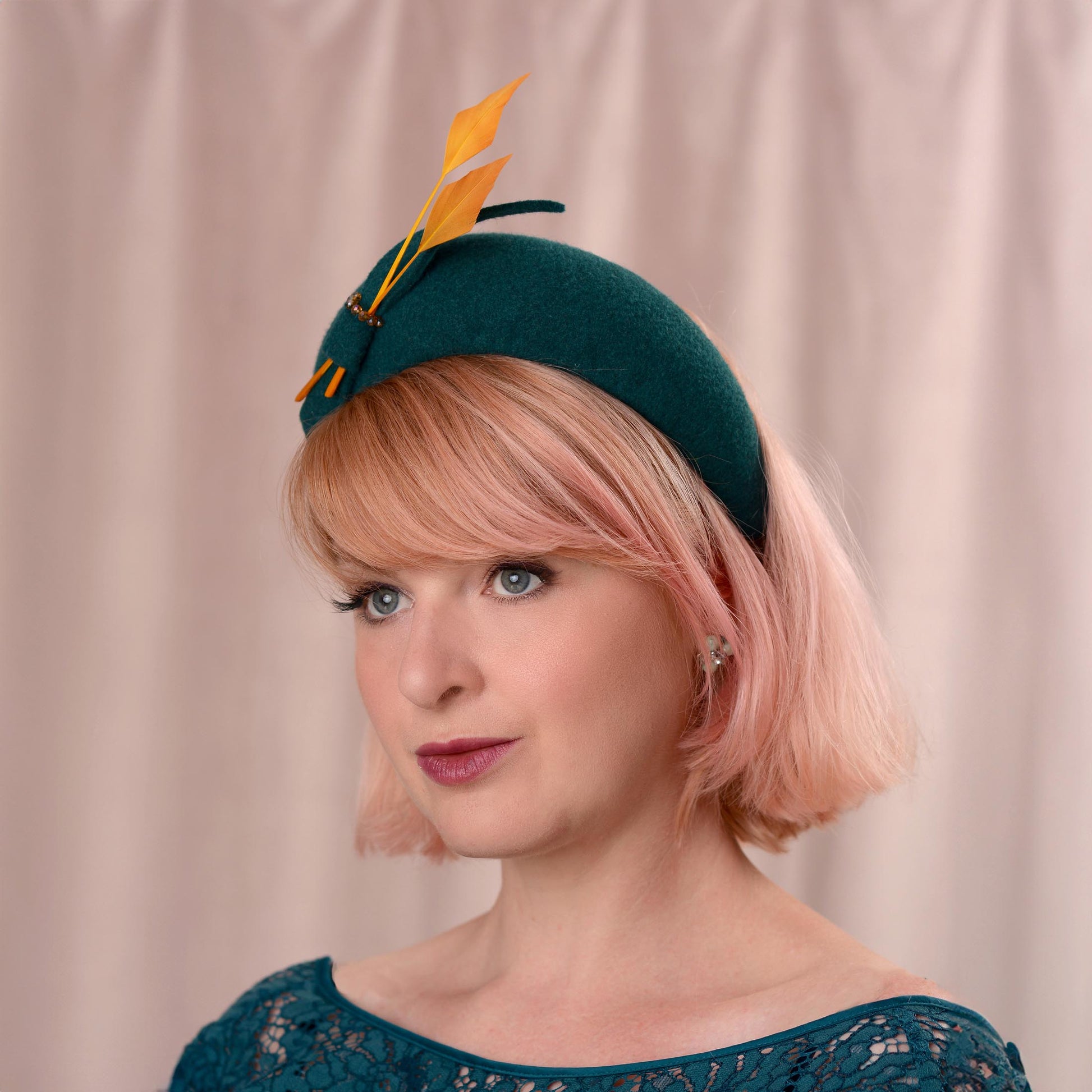 A woman with short pink hair models a dark teal felt fascinator hat decorated with two gold yellow feathers and a small felt bow, set against a soft beige curtain background.
