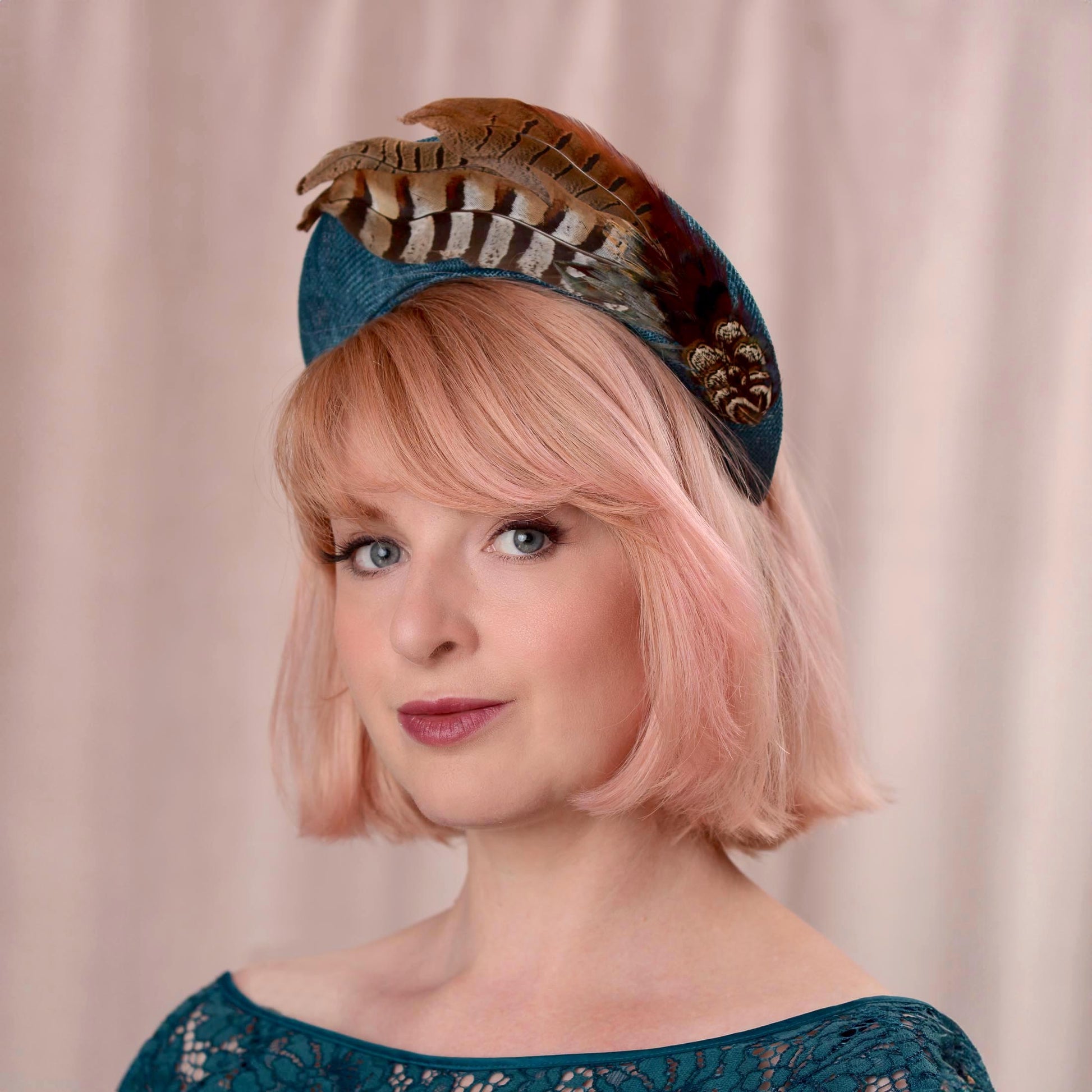 Woman with a pink bob wearing a teal turquoise headband fascinator in a tall elegant style decorated with a plume of pheasant tail feathers with a halo effect. She is wearing a teal dress and standing against a soft pink background