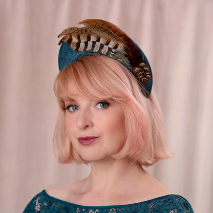 Woman with a pink bob wearing a teal turquoise headband fascinator in a tall elegant style decorated with a plume of pheasant tail feathers with a halo effect. She is wearing a teal dress and standing against a soft pink background