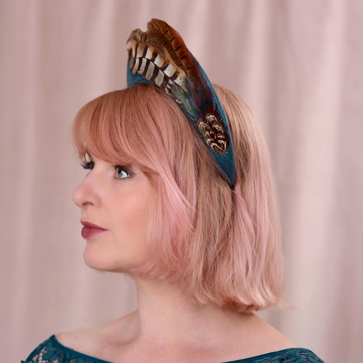 Side view of a woman with a pink bob wearing a teal turquoise wedding headband fascinator in a tall elegant style decorated with a plume of pheasant tail feathers with a halo effect. She is wearing a teal dress and standing against a soft pink background