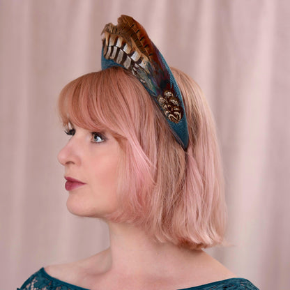 Side view of a woman with a pink bob wearing a teal turquoise wedding headband fascinator in a tall elegant style decorated with a plume of pheasant tail feathers with a halo effect. She is wearing a teal dress and standing against a soft pink background